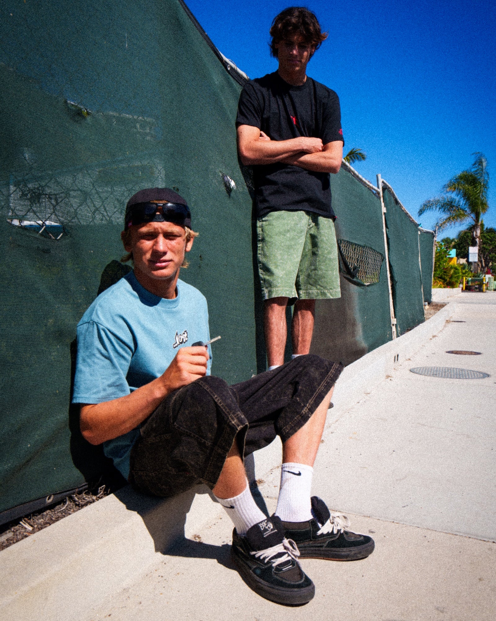 Two men sitting and standing by fence.
